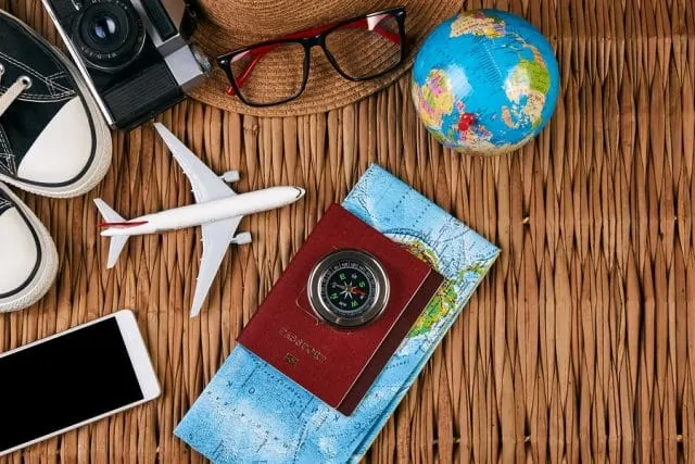 Wicker basket with shoes, camera, hat, toy airplane, toy globe, compass, map, smartphone, and passport on top