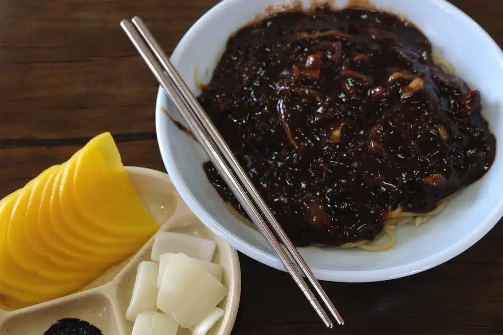 A bowl of black noodle bean sauce. There is also a pair of chopsticks and two side dishes